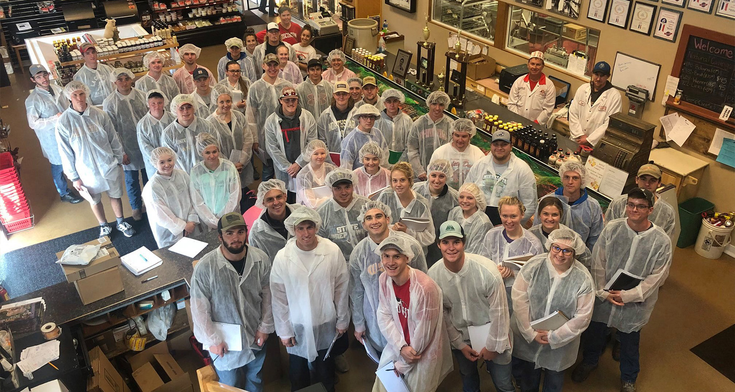 A large group of students stand in hairnets and aprons inside a processing factory