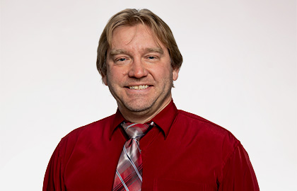 Headshot of Adam Villeneuve, a man with short sandy blonde hair wearing a red button up and red patterned tie