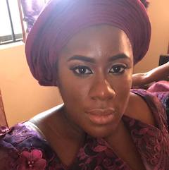 A black woman looks directly at the camera. She is wearing a dark red head wrap and dark red and black top. 