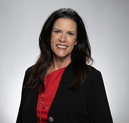 Amy Clark head shot. A woman with long dark hair wearing a red blouse and black blazer in front of a grey background