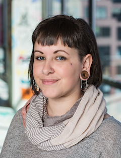 Audrey Williams headshot. White woman with short dark hair, including bangs, wearing a grey sweater and striped scarf and large earrings, smiling straight towards the camera.