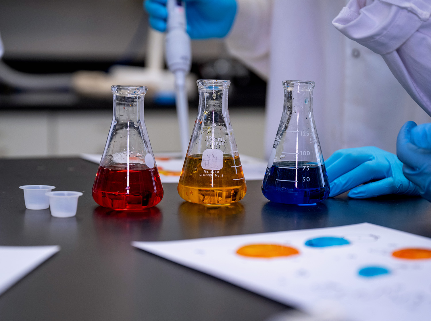 Three flasks sit on a table with red, yellow and blue liquid inside