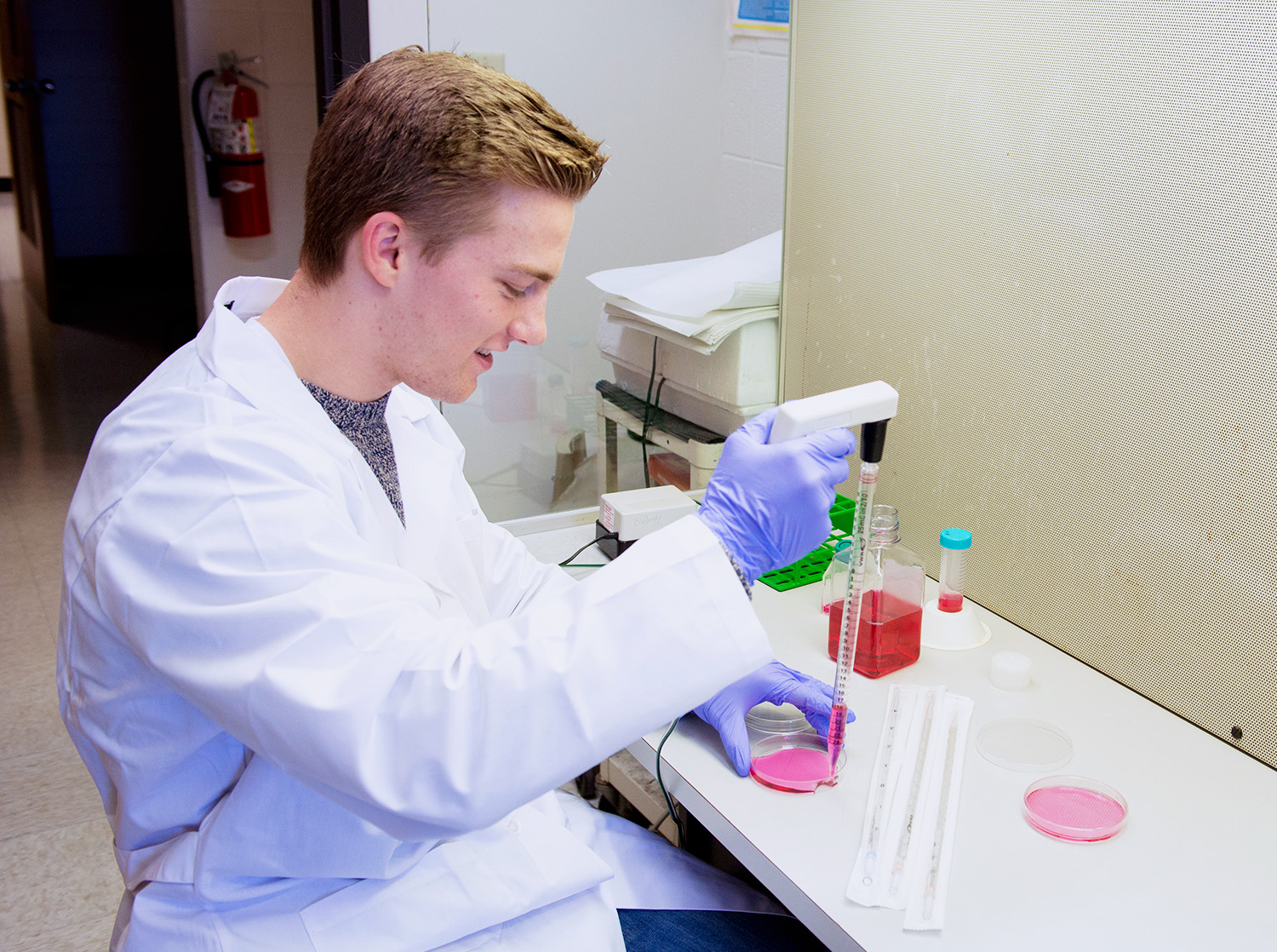 A male student in a white lab coat uses a pipette to transfer liquid