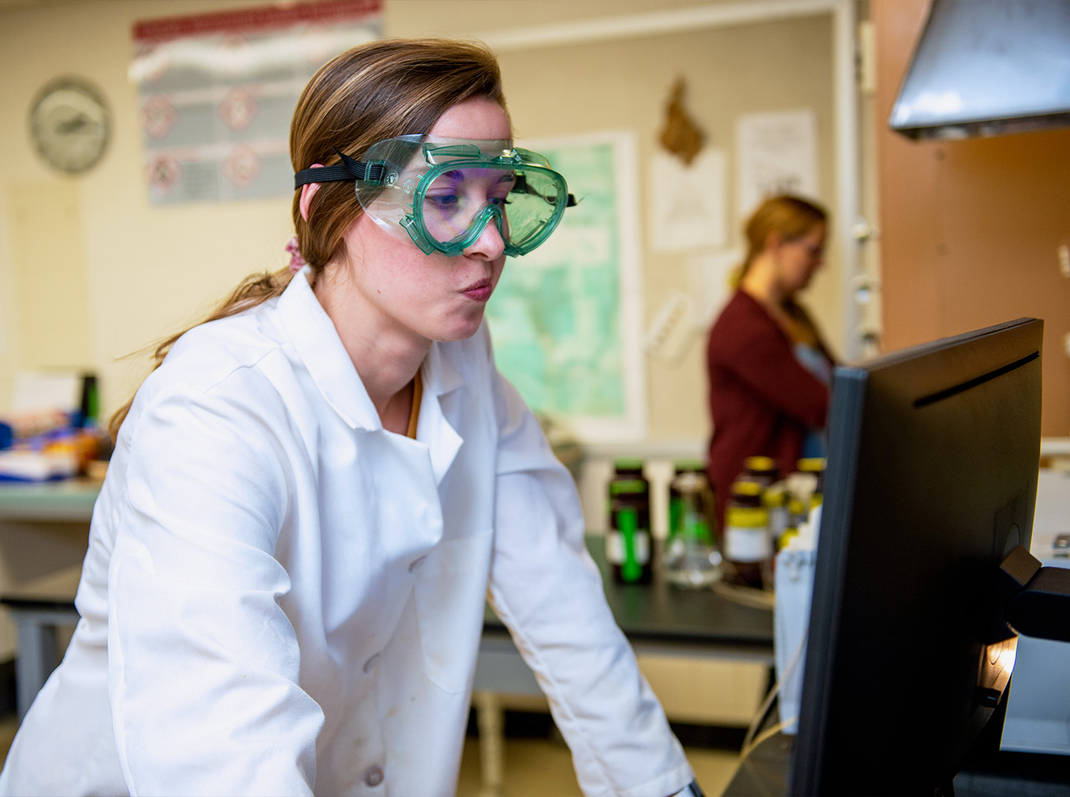 A female student in goggles and a white lab coat works on a computer in the lab