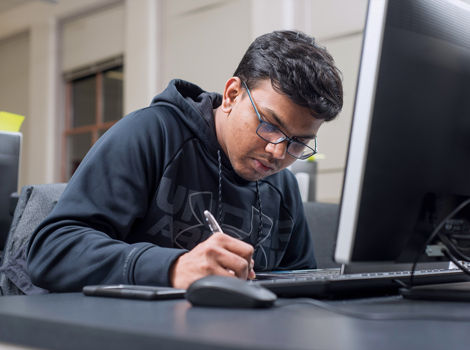 A male student writes in a notebook sitting at a computer desk