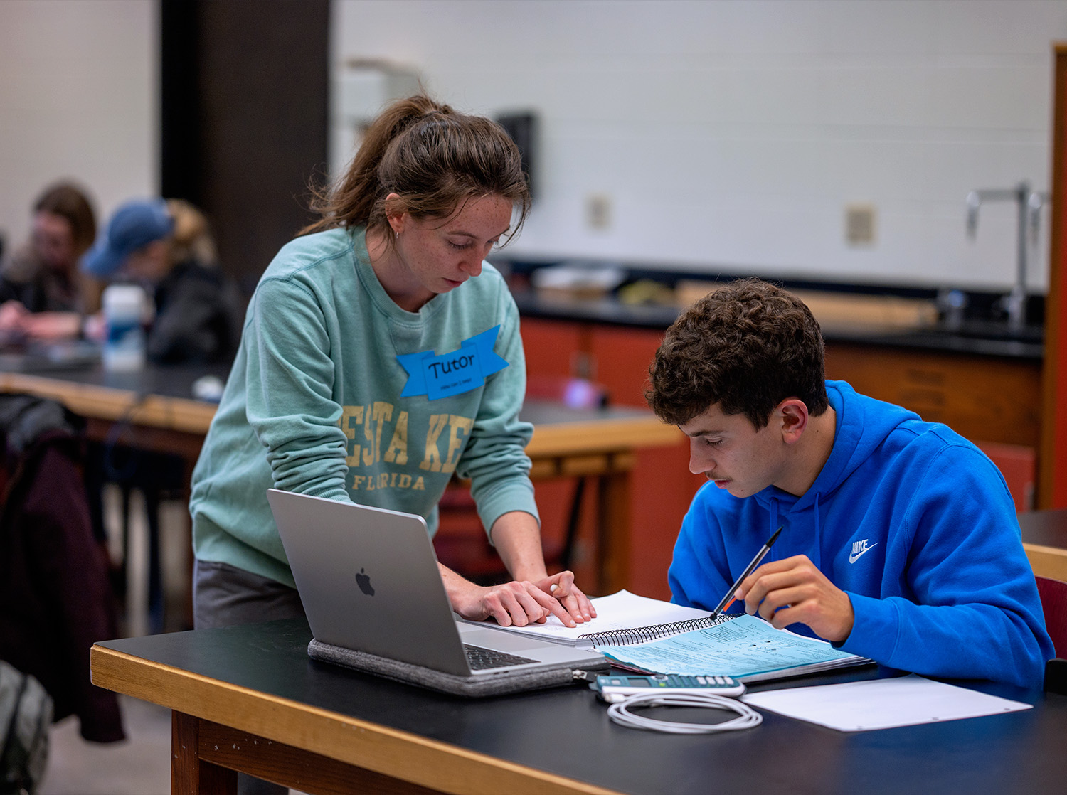 A female student stands over the desk helping a male student with math homework