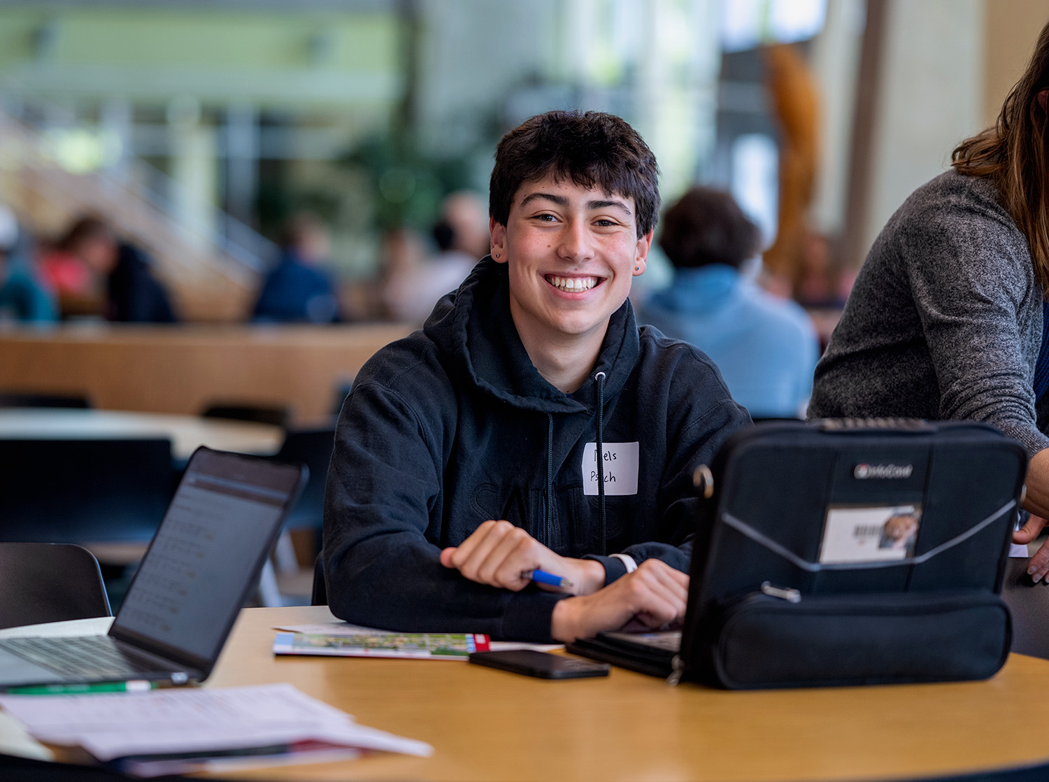A male student smiles at the camera while working on their laptop at a table