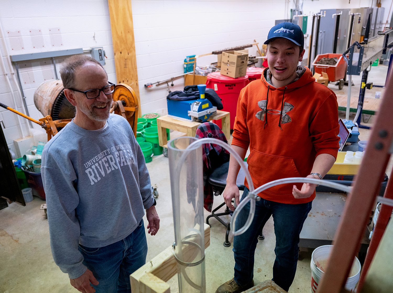 A professor and student stand and observe a large tube during an experiment