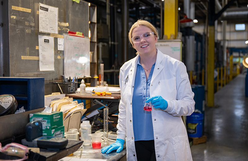 a college age woman with blonde hair wears a blue shirt, blue gloves, a white lab coat and safety goggles. She's holding a beaker of red liquid and standing in front of a lab station