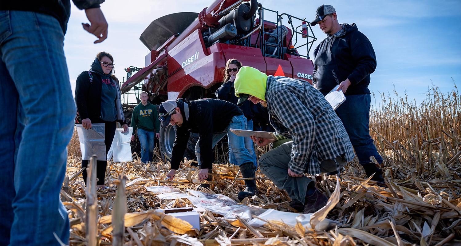 A group of students kneel in a corn field beside a combine