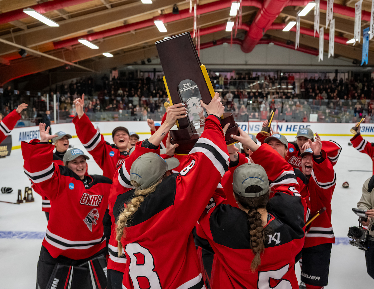 women's hockey team wearing red jerseys lifts national championship trophy up in the air inside the ice arena
