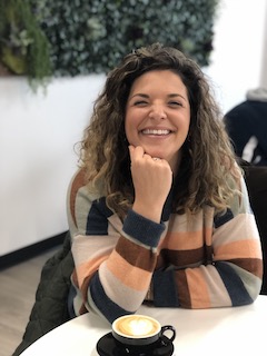 A white woman wearing an orange, brown, tan and blue striped sweater smiles directly at the camera while resting her chin on her fist. She has long wavy dark hair and is sitting at a table with a coffee cup.