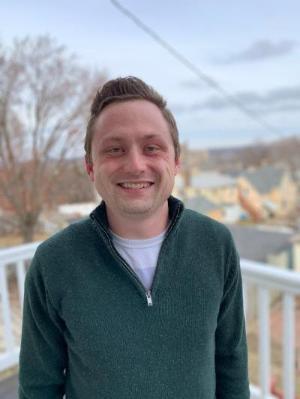 A white man stands on a porch outside and is smiling directly at the camera. He is wearing a green three quarter zip pullover with a white tshirt visible beneath it. He has light hair and no facial hair.