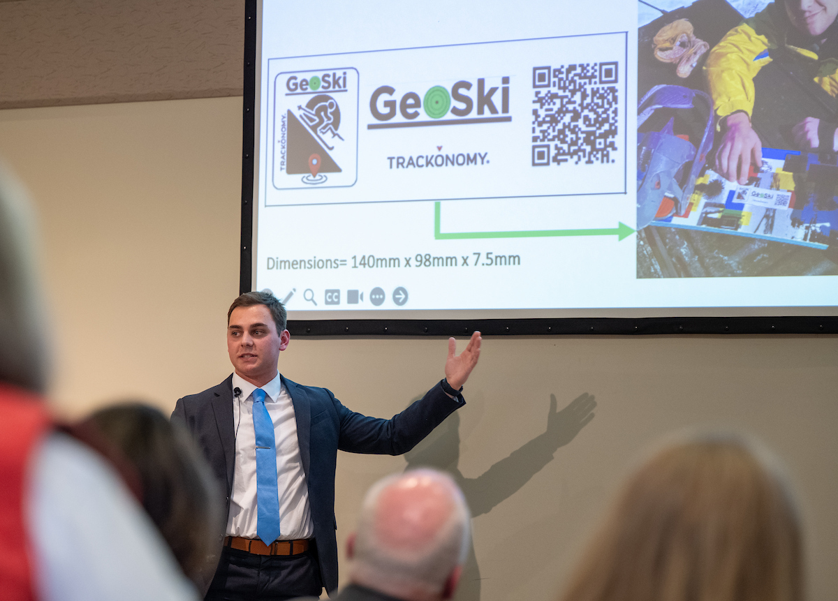 a male presenter stands in front a power point projected on a screen