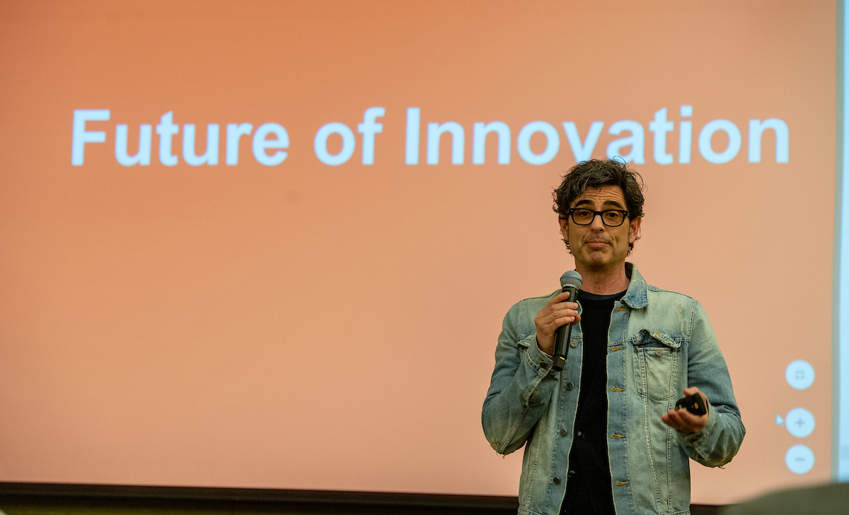 a man wearing a denim jacket and holding a microphone stands in front of an orange screen that says "Future of Innovation"
