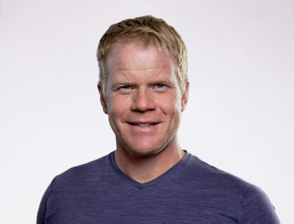 Headshot of Mark Huttermier. A man wearing a dark blue t shirt with short blonde hair