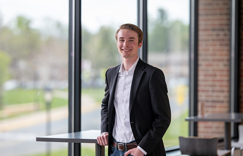 A college aged white male poses in front of a wall of window. He's wearing a white dress shirt and black sport coat.