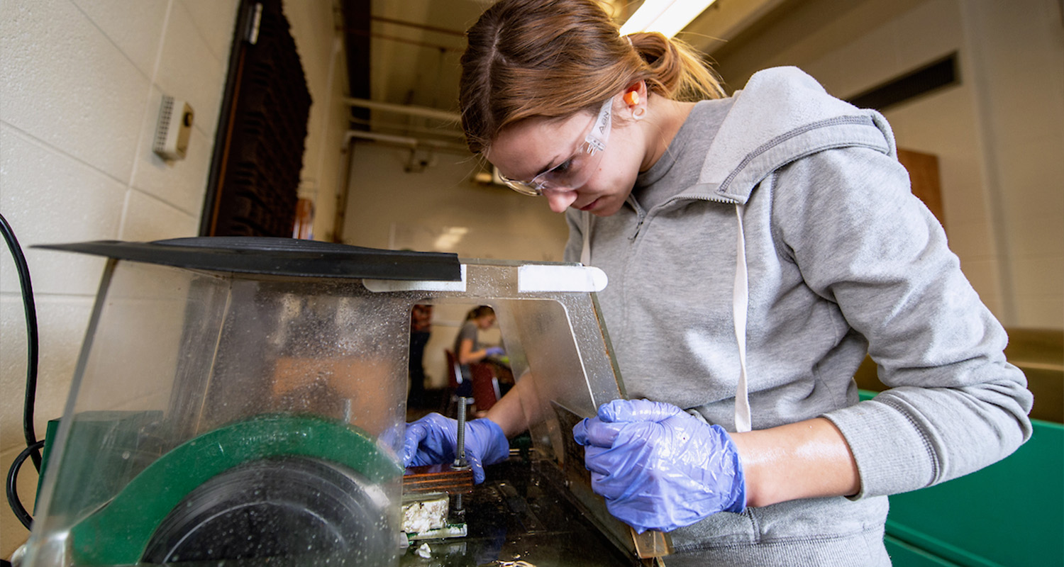 A female student uses a sander to sand down a rock sample to inspect it