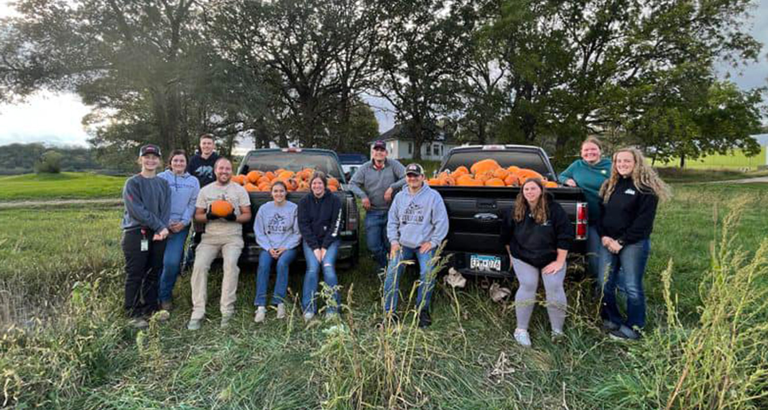 A large group of students sit on the back of two trucks holding multiple orange pumpkins