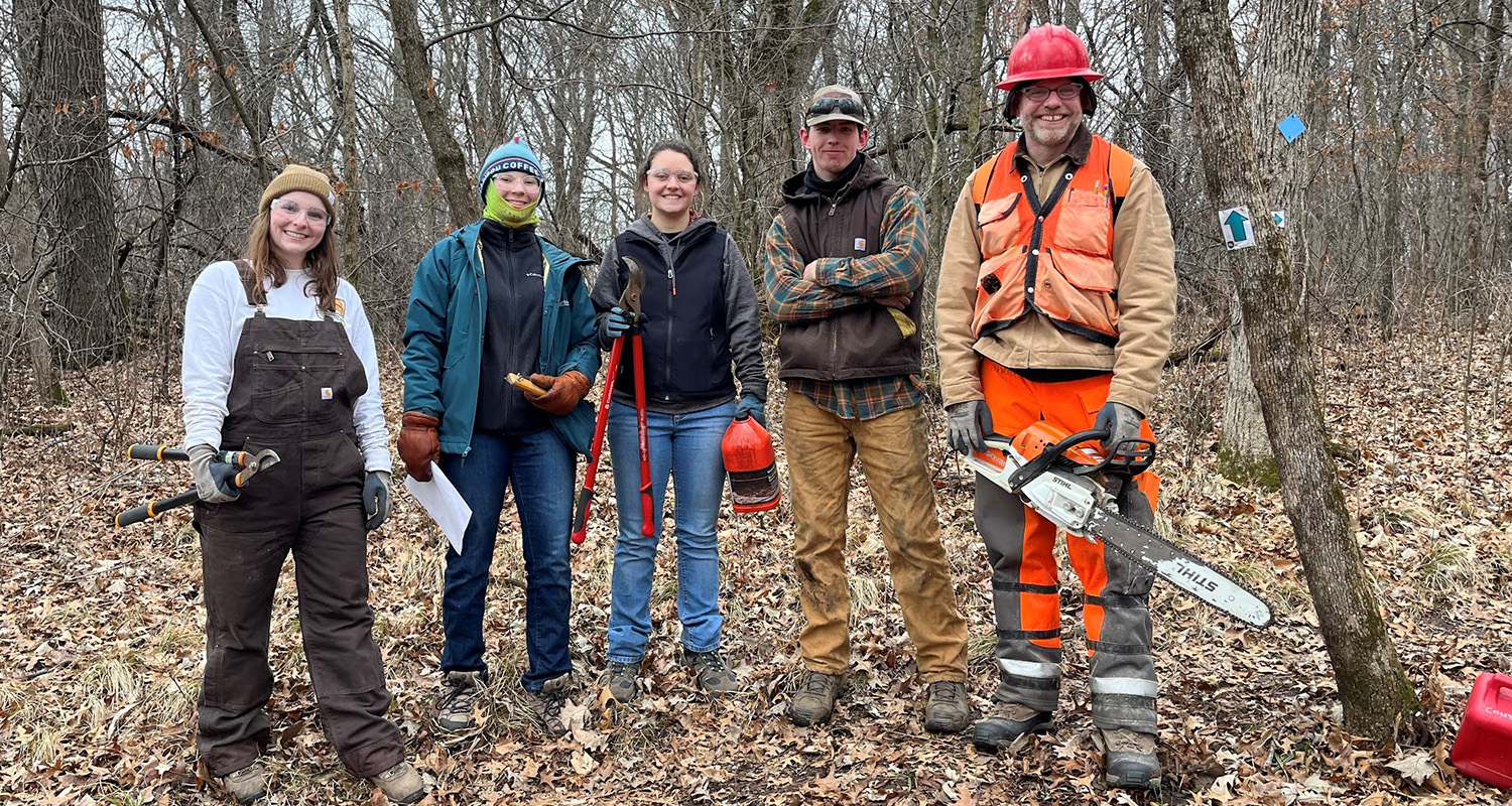 Five people (three females, two males) pose for a photo while standing in a forest, ready to conduct research with a chainsaw and clippers