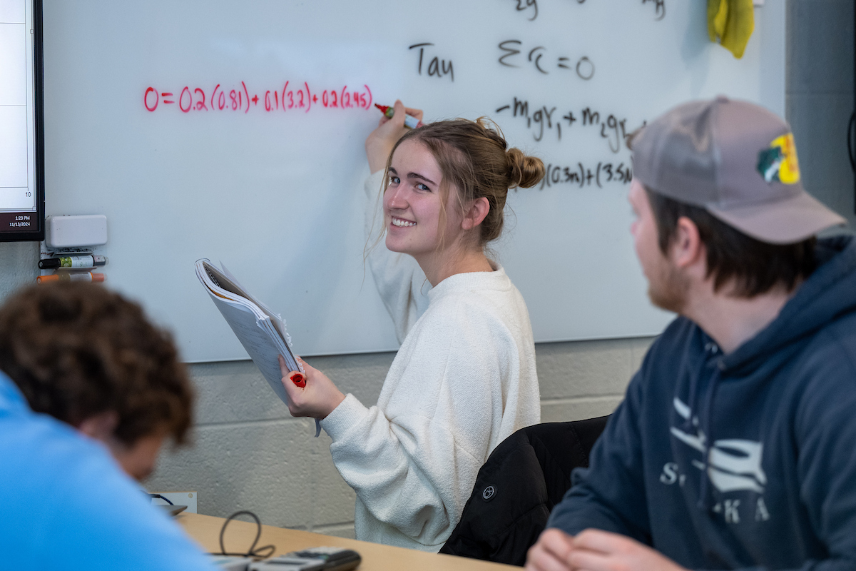 a student stands in front of a whiteboard with physics equations written on it