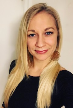 A white woman with long blond hair smiles directly at the camera. She is standing in front of a white wall, wearing a long sleeve black top and large brown earrings. 