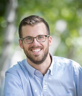 White man wearing a blue button up dress shirt and black glasses is smiling straight toward the camera. He has brown hair and a beard.