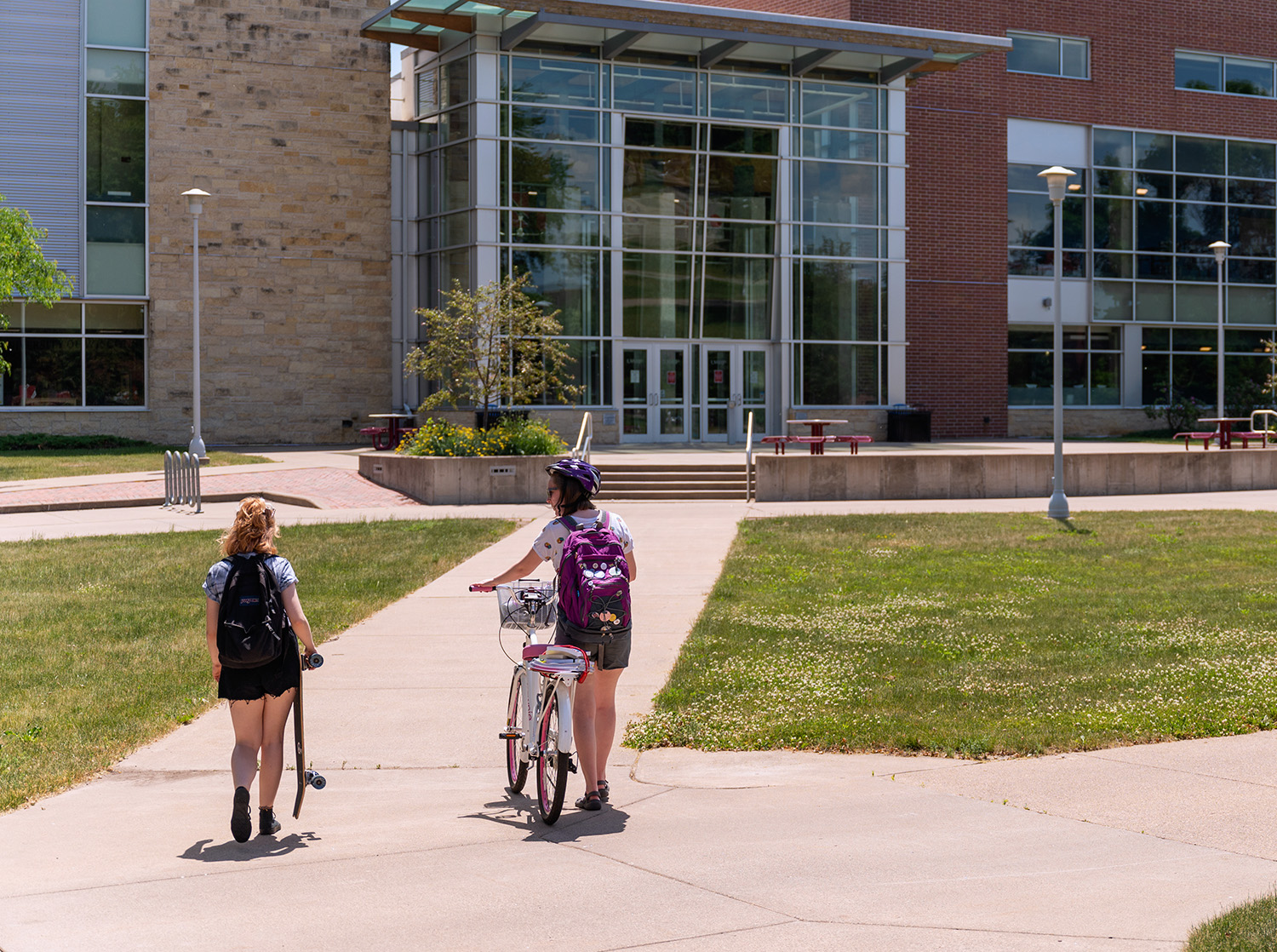 Two female students walk up a path to the University Center. The one one the left holds a skateboard and the one on the right rides a bike with a helmet.