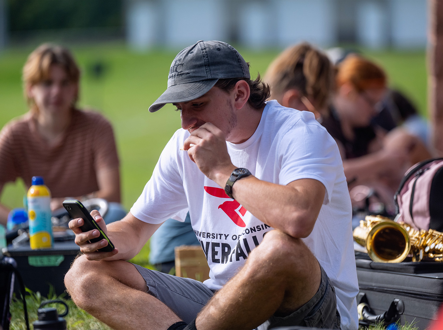 A male student in a baseball hat and UWRF t shirt eats a snack and scrolls on his phone during a break in marching band practice