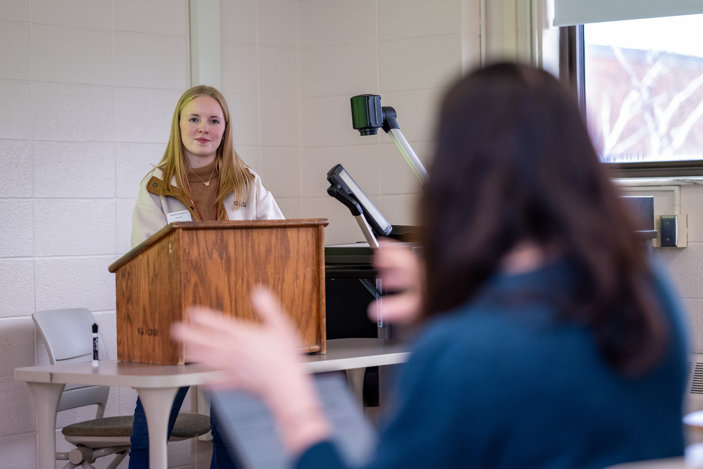 A student stands at a podium in a classroom, listening to someone gesturing in the foreground.