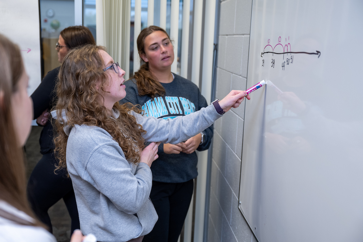 three female students stand at a whiteboard with math equations on it