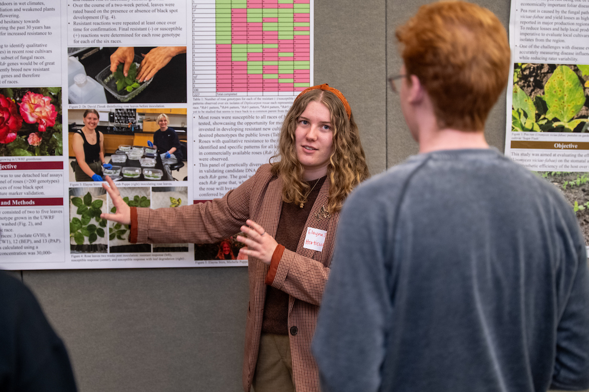 a female student points to a posterboard while she explains the information to an onlooker