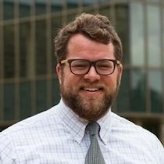 Headshot of Will Erickson, a white man wearing a white button down dress shirt with faint square pattern, green necktie and black glasses looking straight at the camera and smiling. He has dark brown hair and a beard.