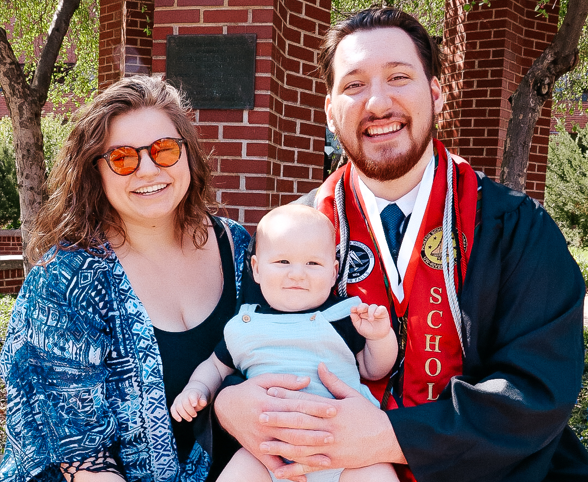 Photo of Greg Peterson in his graduation gown with his wife and baby