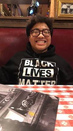 A person sits in a restaurant booth smiling directly at the camera. They are wearing black glasses, a black sweatshirt that says Black Lives Matter in white text and have short dark hair. 