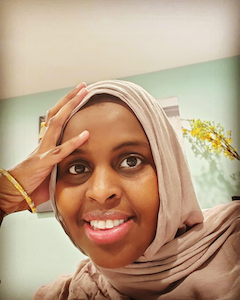 A woman smiles directly at the camera, resting her forehead on her hand. She is wearing a tan headscarf and gold bracelet. 