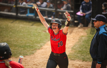 a softball player wearing a red jersey and black pants and helmet raises her arm in celebration as she crosses home plate