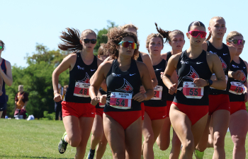 a pack of cross country runners wearing red shorts and black jerseys run on a grass course