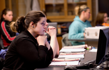 A female student sits at a desk and listens to their professor during class