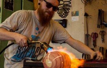 A male student blows flame on a glass piece in the glassblowing studio