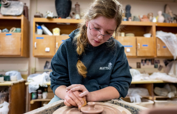 A female student sits at a pottery wheel working on a ceramic piece