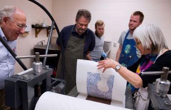 Multiple people stand over a printmaking press while one person pulls a print