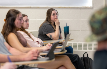 A female student sits at a desk listening to the speaking professor