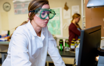 A female student in goggles and a white lab coat works on a computer in the lab