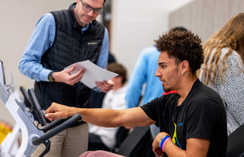 A male student sits on an exercise bike adjusting the machine while the professor watches.