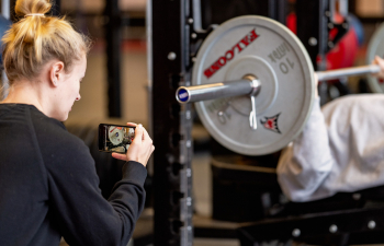 A female student uses their phone to film another student doing a bench press in the Falcon Center