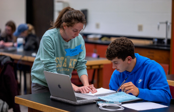 A female student stands over the desk helping a male student with math homework