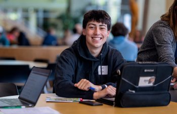 A male student smiles at the camera while working on their laptop at a table