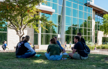 A group of students sit on the grass in a circle in front of the University Center