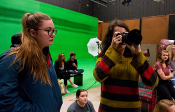 A person looks through a camera lens while standing in front of a green screen